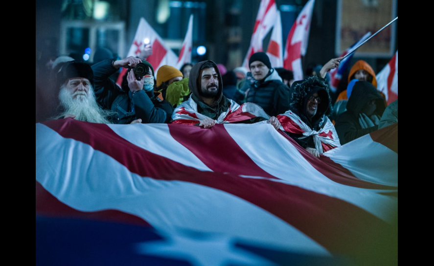 Protesters with a US flag. Screenshot of a photo by Mariam Qashbaia/Publika from February 28, 2026, https://www.facebook.com/photo/?fbid=1694177608648449&set=pcb.1694177725315104